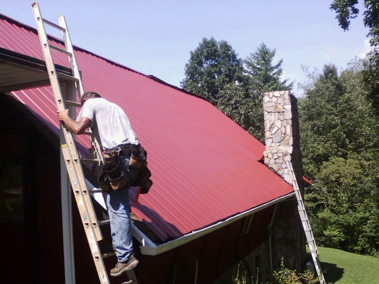 Roofer on a ladder beside a red metal roof and stone chimney — Soffit & Fascia Repair by Roofing Brothers in Ranson